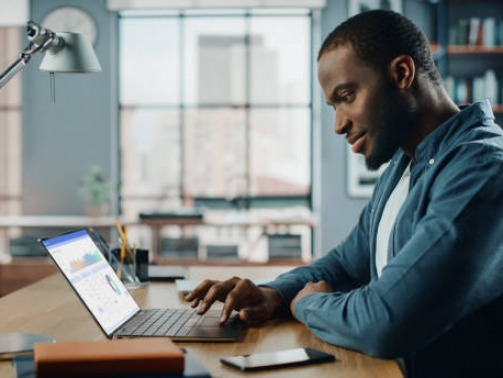 Handsome Black African American Specialist Working on Laptop Computer in Creative Home Living Room. Freelance Male is Doing Market Analysis and Creates Report with Charts for Clients and Employer.